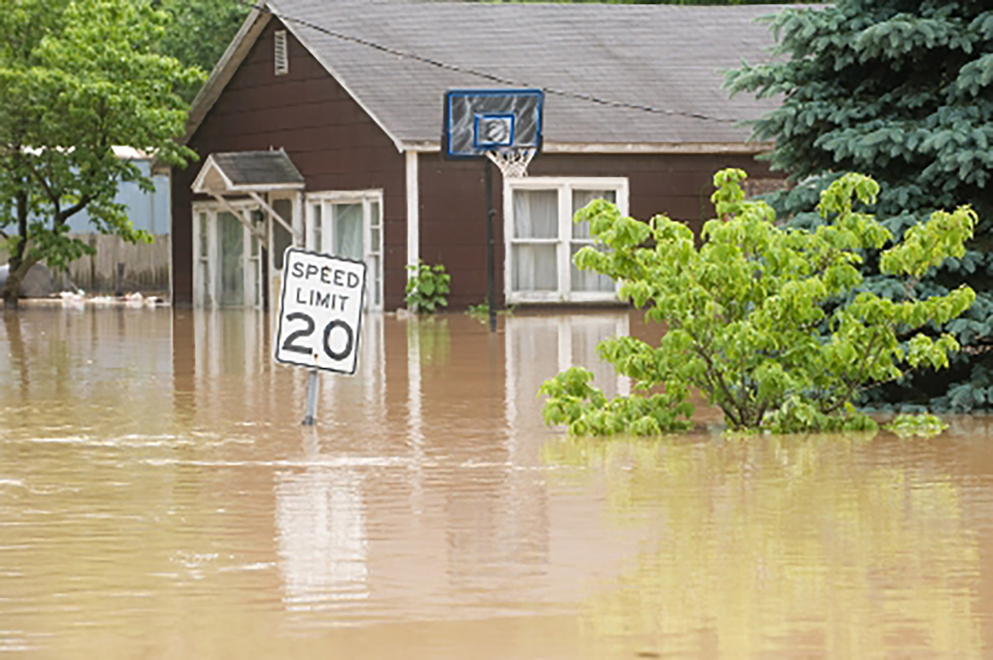 Severe flooding in front of a house up to windows, nearly covering a 20 mph speed limit sign.