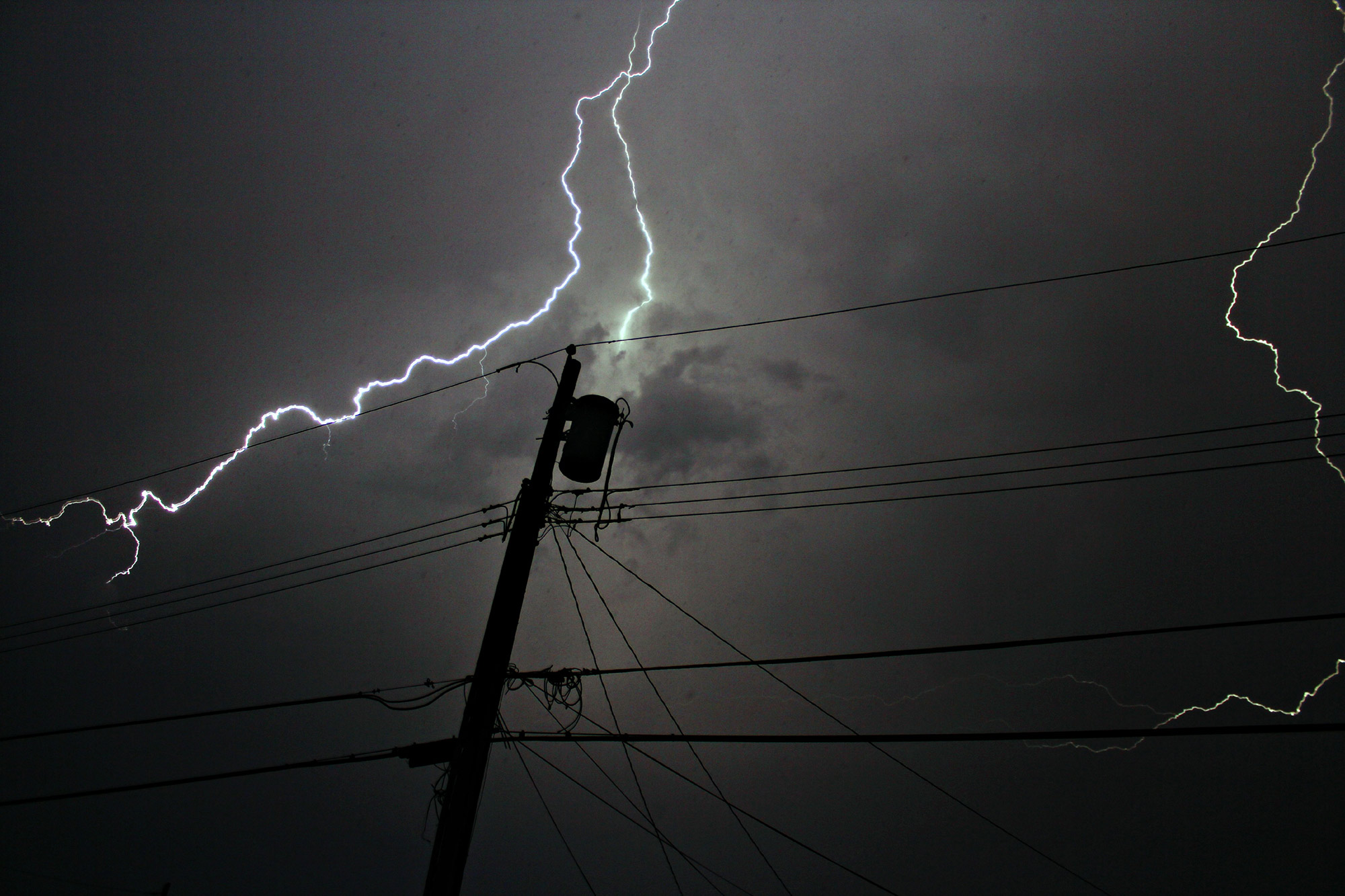 A picture containing dark sky with lightening striking a power line.