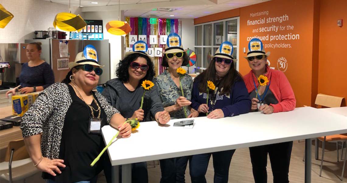 Group of Western National employees posing together wearing matching straw hats and sunglasses and holding sunflowers in lunchroom at Western National's Seattle office.