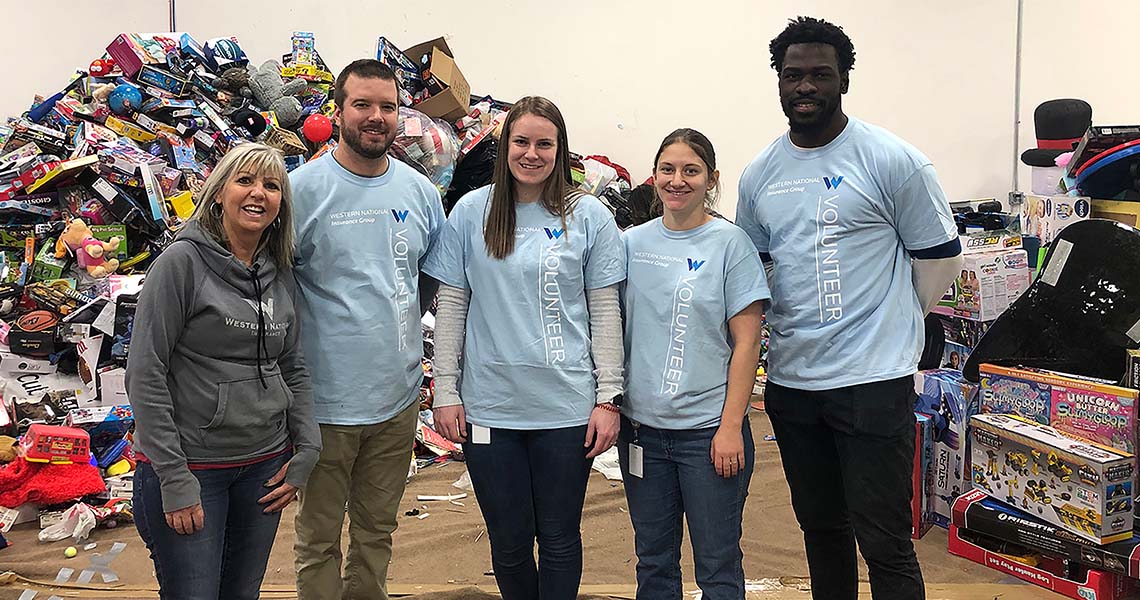 Western National employee volunteers wearing matching Western National branded shirts and posing for a group photo in a warehouse during a volunteer event with large piles of children's toys in the background.